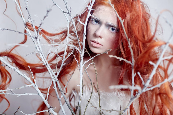 Redhead girl with long hair, a face covered with snow with frost. White eyebrows and eyelashes in frost, a tree branch covered with snow. Snow Queen and winter. Winter makeup woman face, red head. - Fotoğraf, Görsel
