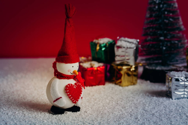 Snowman smiling close to a Christmas tree with gifts. Snowy floor and red background. - Фото, изображение