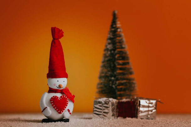 Snowman, christmas tree and gifts over snowy floor and orange background. Christmas still life. - Photo, image