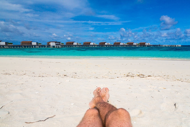 Fondo de viaje con dos patas masculinas en una playa con un mar turquesa y villas de agua en el fondo
 - Foto, Imagen
