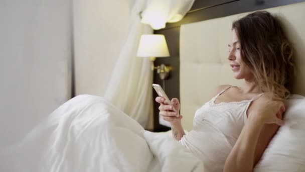 Young beautiful woman lying on bed and checking social networks on a smartphone - Кадры, видео