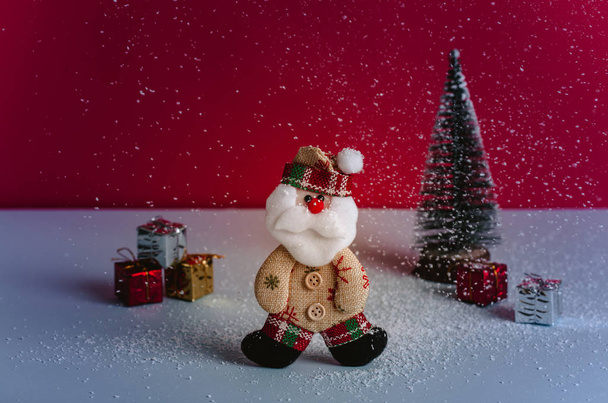 Cute Santa felt doll between a gifts pile and a Christmas tree while snowing. Red background and beautiful light. - 写真・画像
