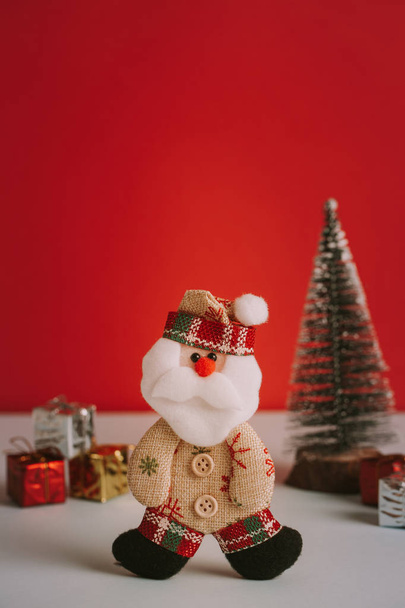 Cute alternative Santa toy with red background and gifts and Christmas tree. Vertical shot and copy space. - 写真・画像