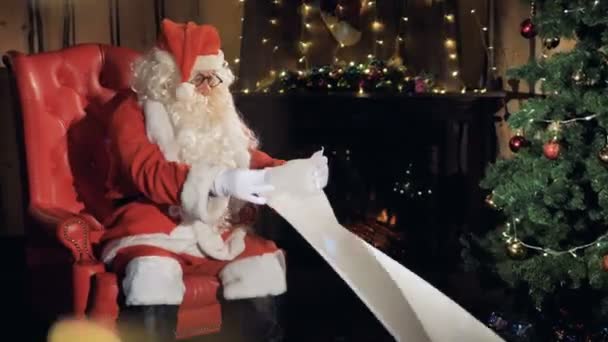 Santa Claus sits in his chair in front of a Christmas tree reading new year gift list. - Πλάνα, βίντεο
