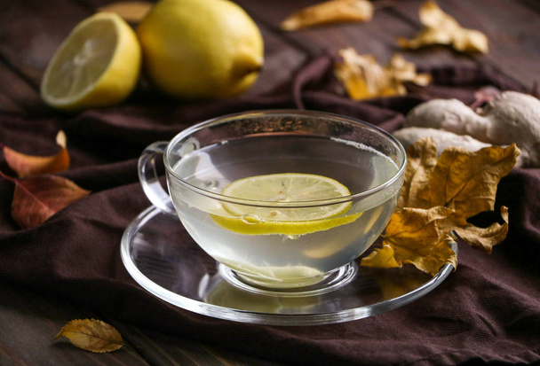 Jedna skleněná šálek užitečných horký čaj s citronem a pikantní zázvor, který chrání proti chřipce a nachlazení, suché podzimní listí v hnědé textilní ubrousek na hnědý dřevěný stůl - Fotografie, Obrázek