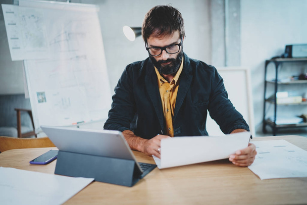 Handsome bearded male architect wearing eye glasses working on a paper plan at his desk. Professional experienced engineer constructionist developer.Horizontal.Blurred background. - Photo, Image