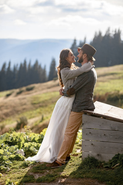 Jonggehuwden staan, glimlach en knuffel op achtergrond van bergen. - Foto, afbeelding