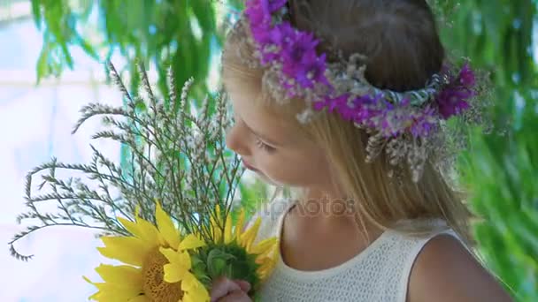 Little blonde with flowers crown and flowers in hands smiles at camera 4K - Video