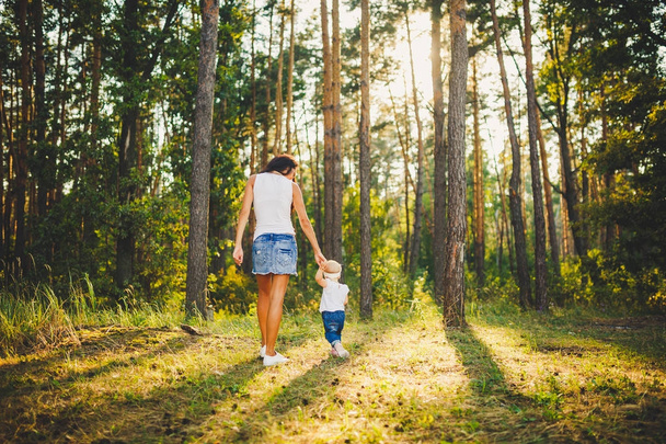 美しく、スタイリッシュな母は、背の高い針葉樹の背景に公園で誕生から 1 年の女の子の手を導きます。家族の価値観のお母さん子を歩くことを学ぶ - 写真・画像