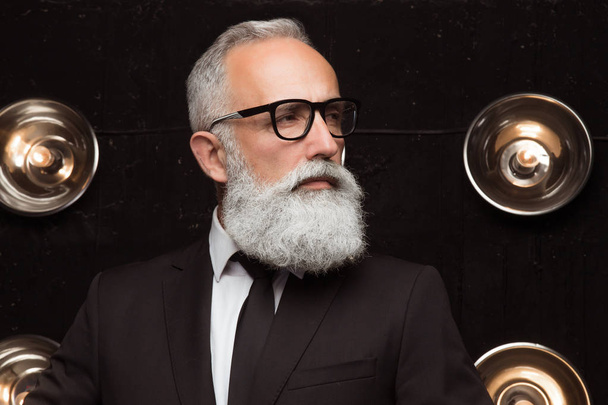 Pensive man in fashion glasses look away. Styling old man with a long white beard posing on the dark background in modern strict suit. Portrait of modern Santa Claus - Photo, Image