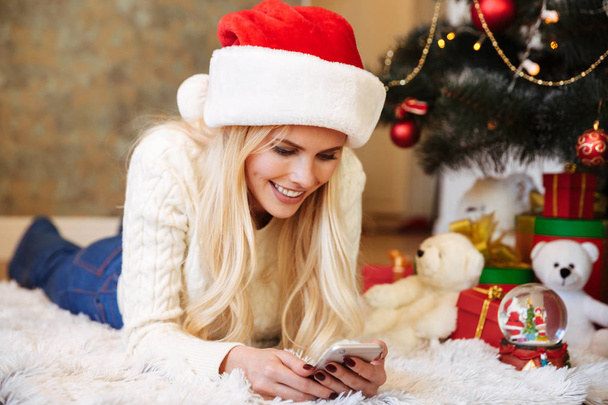 Smiling blonde woman in Santa's hat checking news on smartphone  - 写真・画像