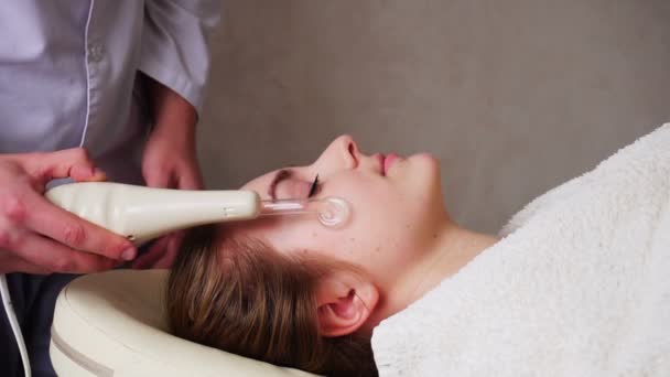 Hombre experto cosmético demuestra el trabajo de dispositivo de masaje para la cara en el fondo de la niña pacientes, que se encuentra en el sofá en la oficina de salones
. - Metraje, vídeo