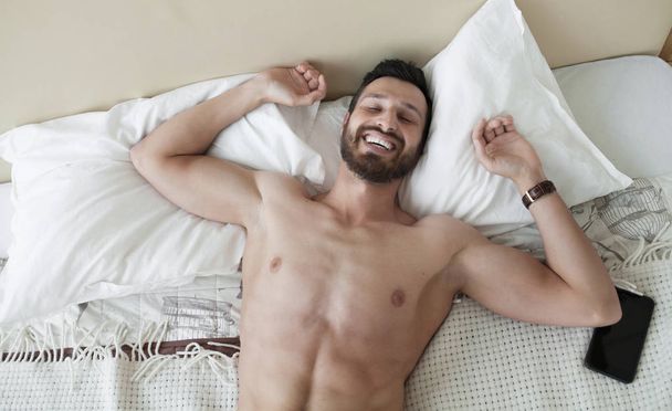 Carefree guy enjoying new day. Sexy, happy bearded man in bed. - 写真・画像