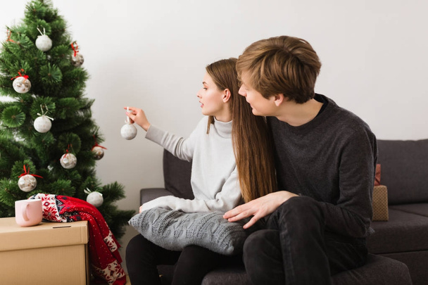 Portrait of beautiful young couple sitting on sofa at home and decorating Christmas tree together - Foto, Imagem