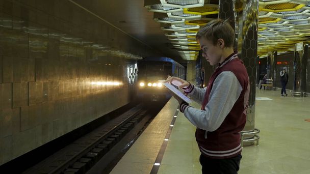 Člověk pomocí touchpadu nebo smartphone, když on čeká na vlak ve stanici metra. Osamělý mladý muž s smartphone střílel z profilu na stanici metra s rozmazaný jedoucího vlaku v pozadí. - Fotografie, Obrázek