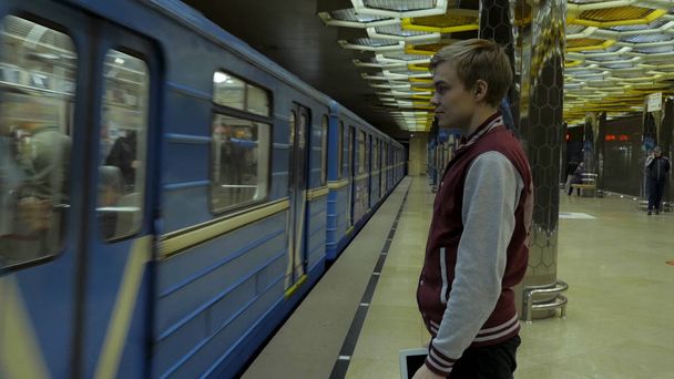 彼は地下鉄の駅で電車を待つときにタッチパッドやスマート フォンを使用している人。スマート フォンで孤独な若い男がプロファイルからぼやけて背景で動いている電車と地下鉄の駅で撮影. - 写真・画像