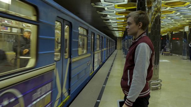 彼は地下鉄の駅で電車を待つときにタッチパッドやスマート フォンを使用している人。スマート フォンで孤独な若い男がプロファイルからぼやけて背景で動いている電車と地下鉄の駅で撮影. - 写真・画像