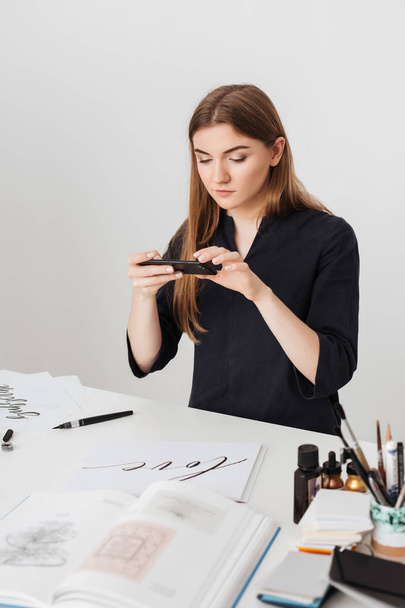 Portrait of young beautiful lady sitting at the white desk and taking photos of cool notes on her cellphone isolated - Foto, Imagen
