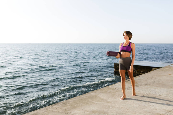 Młoda kobieta uśmiechający się ciemne włosy krótkie, w ręku trzyma matę do jogi podczas spaceru przez morze. Piękna Pani w sportowy top i spodenki stojący i sennie patrząc na bok, z widokiem na morze na tle - Zdjęcie, obraz