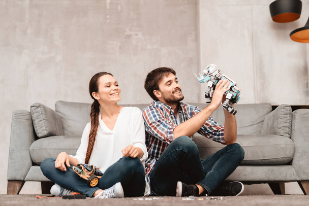 A young couple is sitting on the floor in the living room. The guy is holding a robot. The girl sits next to the guy and pulls to the robot. - Foto, imagen