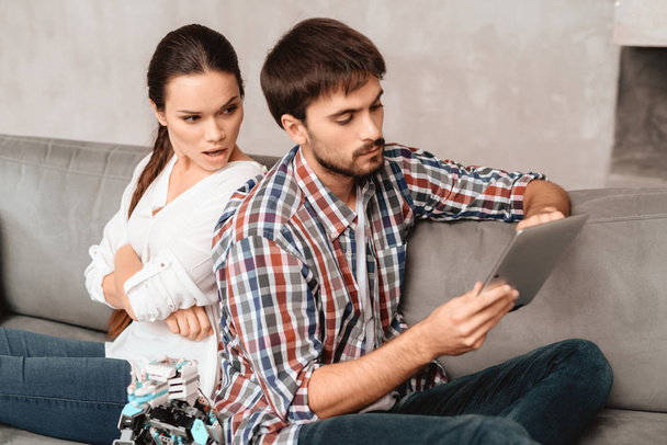 Young couple in the living room. The guy is holding a robot. The girl next to him, and she is very unhappy that the guy ignores her. They are sitting on the gray couch in the living room. - 写真・画像