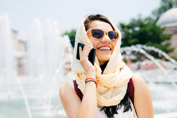 happy swarthy arab woman in headscarf, wearing sunglasses talking on the phone and smiling - Foto, afbeelding
