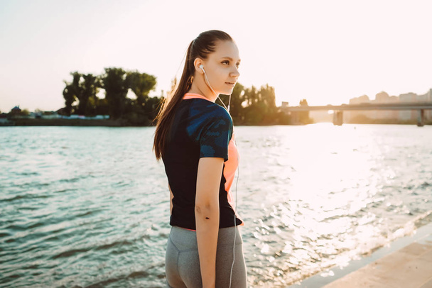 athletic girl in sports clothes walking by the river, listening to music on headphones, sunset - Valokuva, kuva