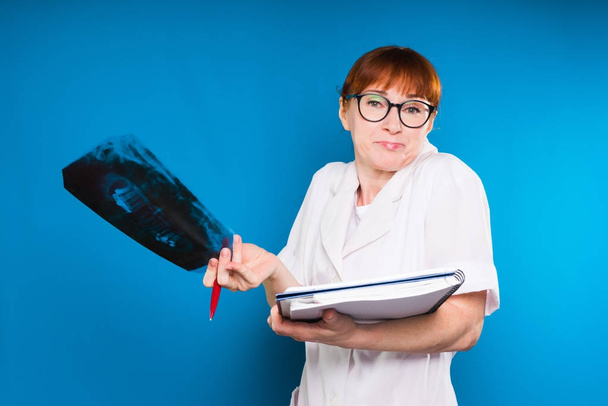 adult red-haired woman doctor wearing glasses holds an x-ray of teeth and a notebook, does not know what to do - Фото, изображение