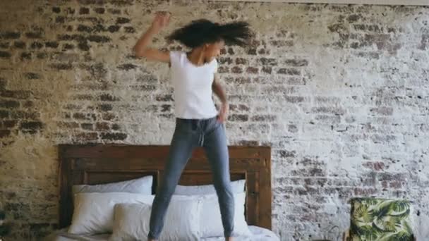 Curly mixed race young joyful girl have fun dancing on bed at home - 映像、動画