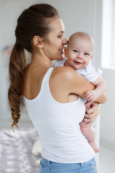 A happy young woman,a brunette with thick hair braided in a thick braid,a make-up and a pleasant smile,dressed in blue trousers and a white jersey, holds a newborn son in her arms,a boy with short,fluffy hair in a white T-shirt,a photo in the bedroom - Photo, Image