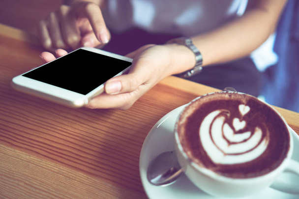 Młoda dziewczyna trzymając telefon biały ma pusty ekran w kawiarni, i kawa na tabelę, aby uczynić życie bardziej relaksujący. Zastosowanie technologii dla szczęśliwego życia. - Zdjęcie, obraz