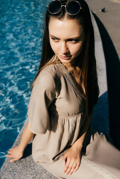  charming girl in a stylish beige dress sits by the pool with blue water, enjoys the sun - Fotó, kép