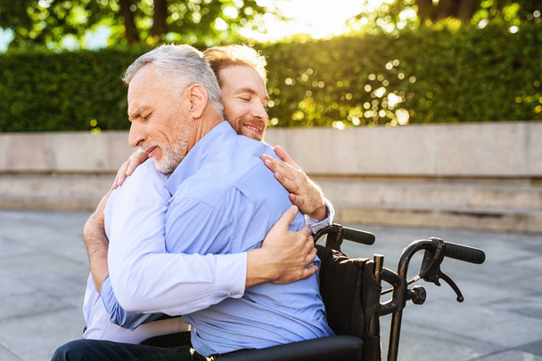 To the old man who sits in the park on a wheelchair came his son. He is hugging the old man - 写真・画像
