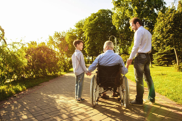 Vanha mies pyörätuolissa, poika ja pojanpoika kävelevät puistossa.
 - Valokuva, kuva