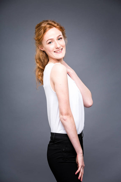 portrait of a young redhaired beautiful girl in the studio on a gray isolated background. A woman is standing with her arms folded and smiling in a white shirt with a short sleeve. Business concept - Zdjęcie, obraz