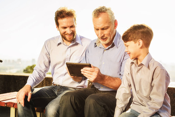 The old man his son and grandson are sitting on the bench watching something on the tablet - 写真・画像