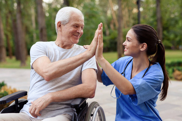 A nurse and an old man in a wheelchair high five in a summer park - Foto, immagini