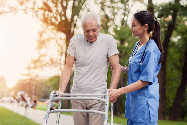 The old man walks through the park with the help of adult walkers. The nurse supports him - Photo, Image
