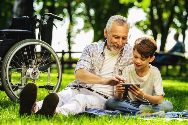 The old man and his grandson are sitting on a picnic and are looking at something on the smartphone - Photo, image