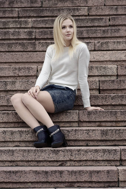 ein schönes Mädchen in Jeansrock und weißem Strickpullover sitzt auf den Stufen. - Foto, Bild