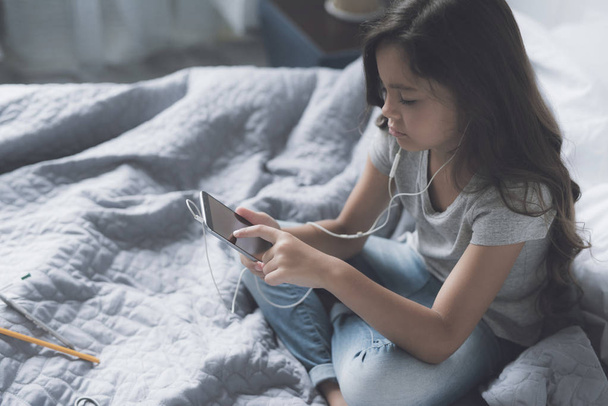 Little black-haired girl sitting on the bed cross-legged and listening to music on a gray smartphone in the morning - Photo, image