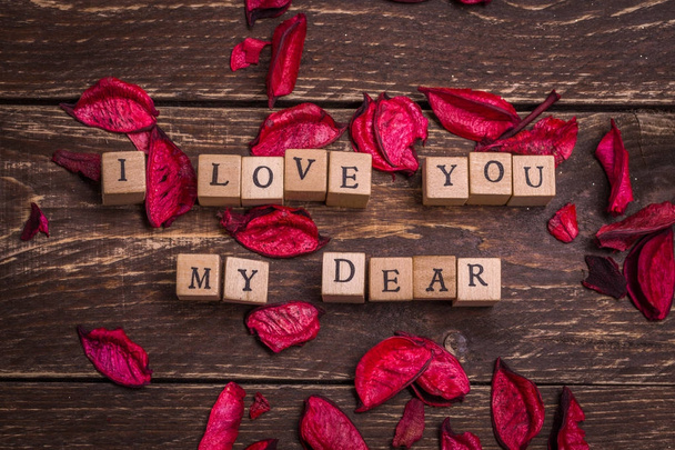 I love you my dear. Valentines card. Small wooden letters on wood cubes standing on dark rustic vintage background. Red and violet pink petals around at the sides. Greetings postage anonymous message. - Foto, Bild