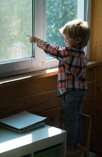Мальчик показывает палец на что-то снаружи окна на улице. Красивая блондинка стоит на стуле и смотрит из дома в окно. Детство, ожидание и радость
 - Фото, изображение