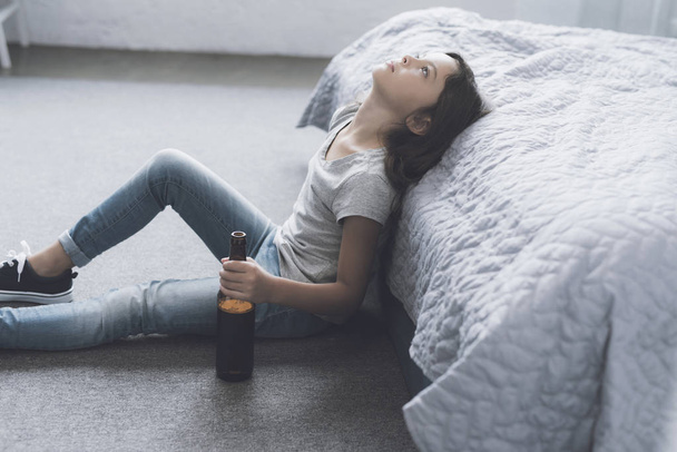 Girl sitting on the floor leaning on a gray bed and looking at the ceiling, holding in her hand an open bottle of beer - Photo, image