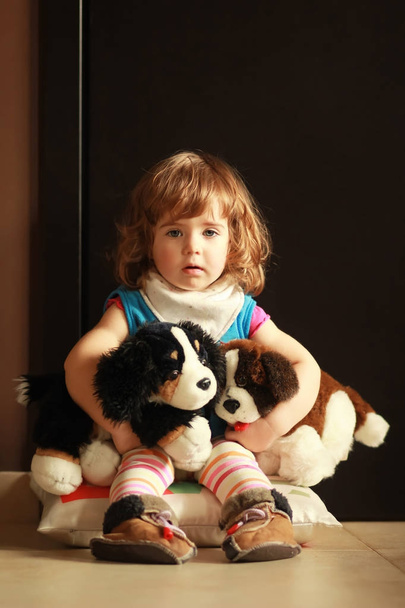 Portrait of a cute one year old girl sitting at home on a pillow on the floor and hugging her favourite toys - dogs.  - Photo, Image