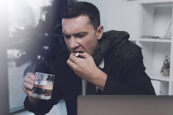 A sick man is sitting at his workplace in the office. He has a pill and a cup of water in his hands - Photo, image