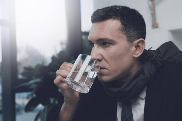 A sick man is sitting at his workplace in the office. He holds a cup of water in his hands - Zdjęcie, obraz