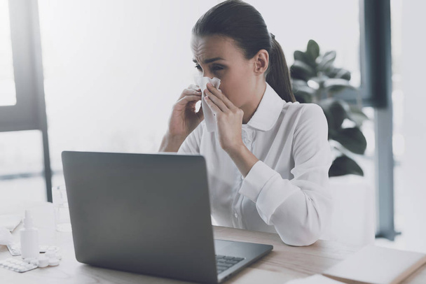 Sick woman sitting at her workplace in the office. A woman blows her nose in the workplace - Foto, Imagem