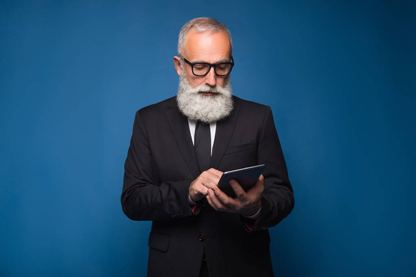 Concentrate bearded senior using internet on a tablet and working in a comfortable way. Serious man in a glasses and modern strict suit - Photo, Image