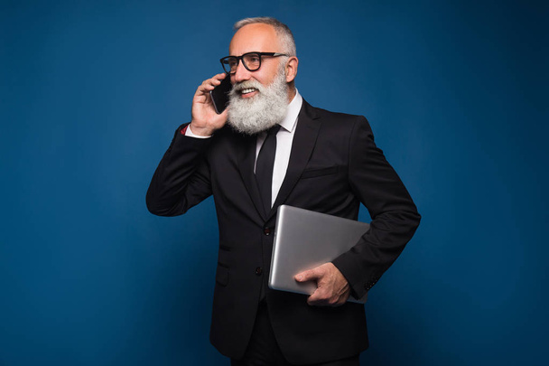 Ocupado barbudo sênior e homem de negócios ficar com notebook e telefone celular isolado em um fundo azul
.  - Foto, Imagem